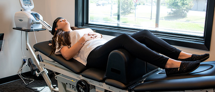 A woman relaxing during a spinal decompression therapy session in a chiropractic clinic. She is lying on a treatment table with her eyes closed, while a spinal decompression machine is positioned nearby, with a view of the outdoors through the window.