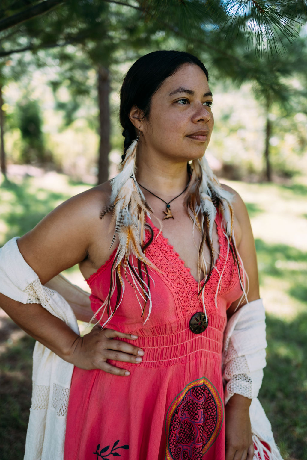Ksandra Roehr in a coral dress with feather earrings, standing under pine trees