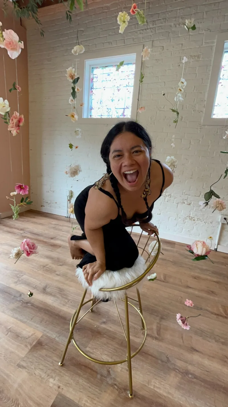 Ksandra laughing, perched on a gold stool among hanging flowers