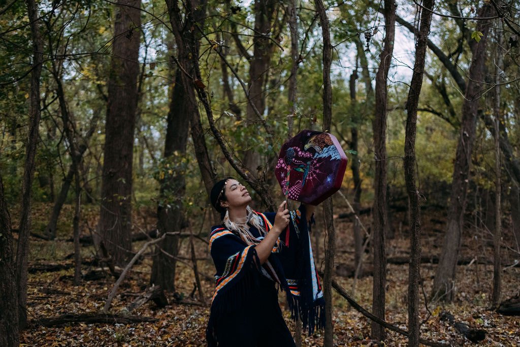 Ksandra Roehr lifting a painted ceremonial drum in an autumn forest