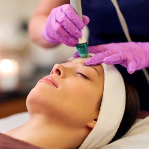 A HydraFacial handpiece being passed over a client&rsquo;s forehead while she lies back with her eyes closed