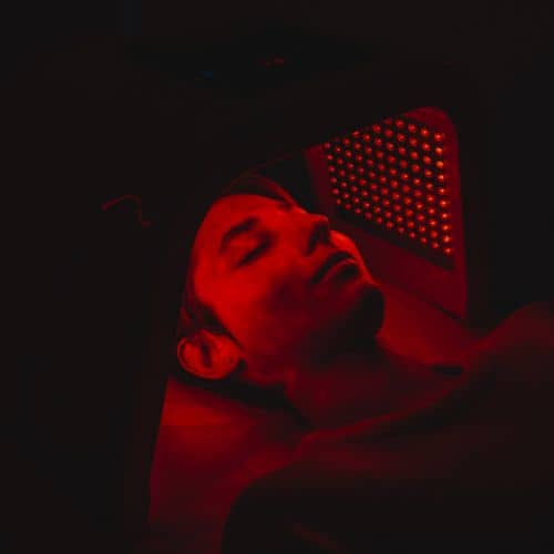 A client lying back with eyes closed under a panel of red light, the light washing over the face and chest