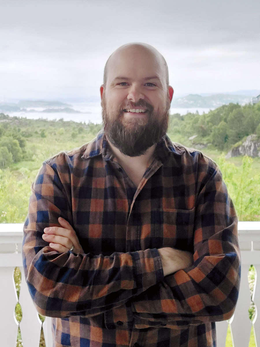 Travis D. Peterson, bald man with beard wearing a brown flannel shirt stands smiling with arms crossed in front of a white railing and a green valley behind him.