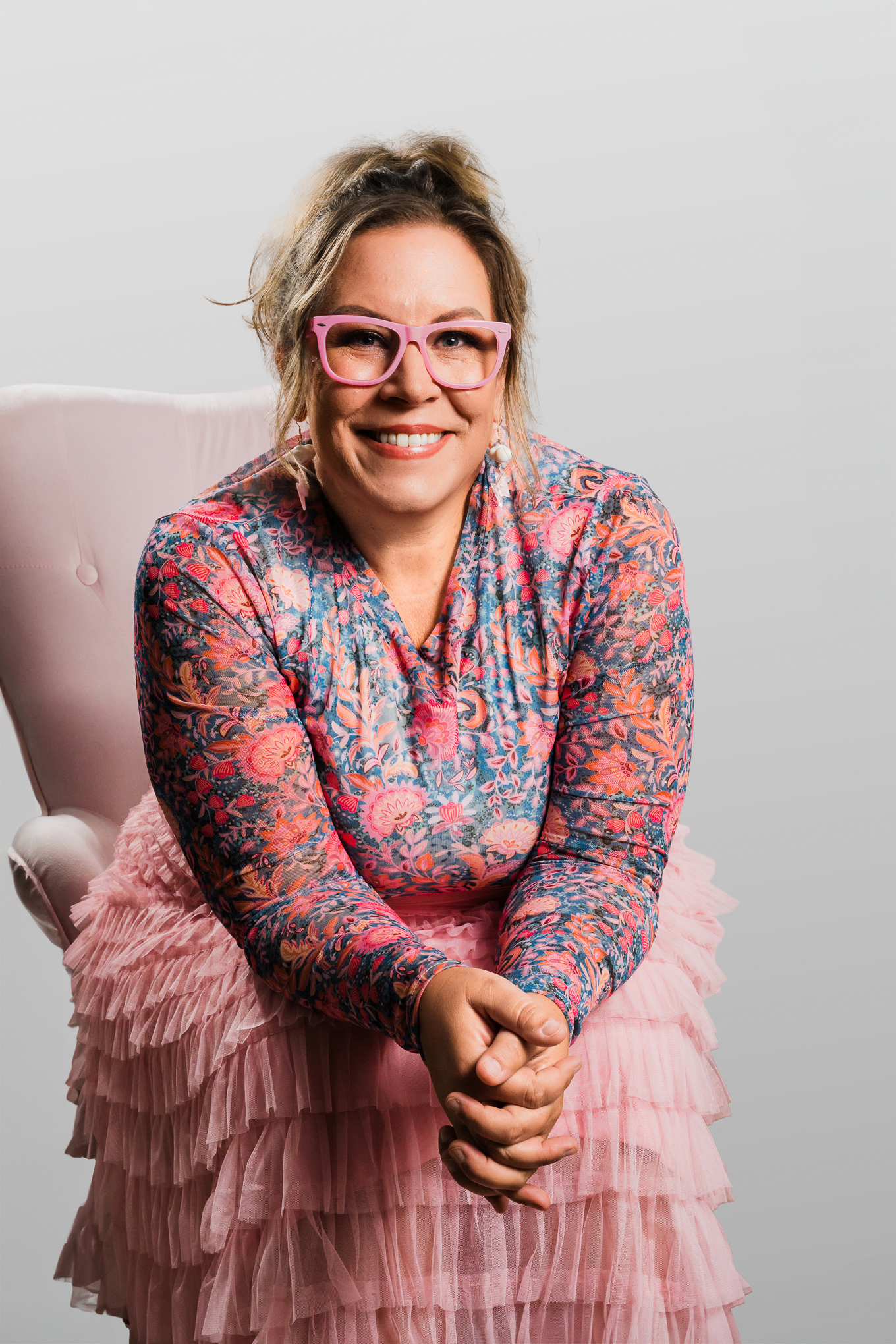 Wendi Fredregill seated and smiling in pink glasses, floral top, and tulle skirt — founder of Hey Wendi, a clarity practice for overwhelmed women