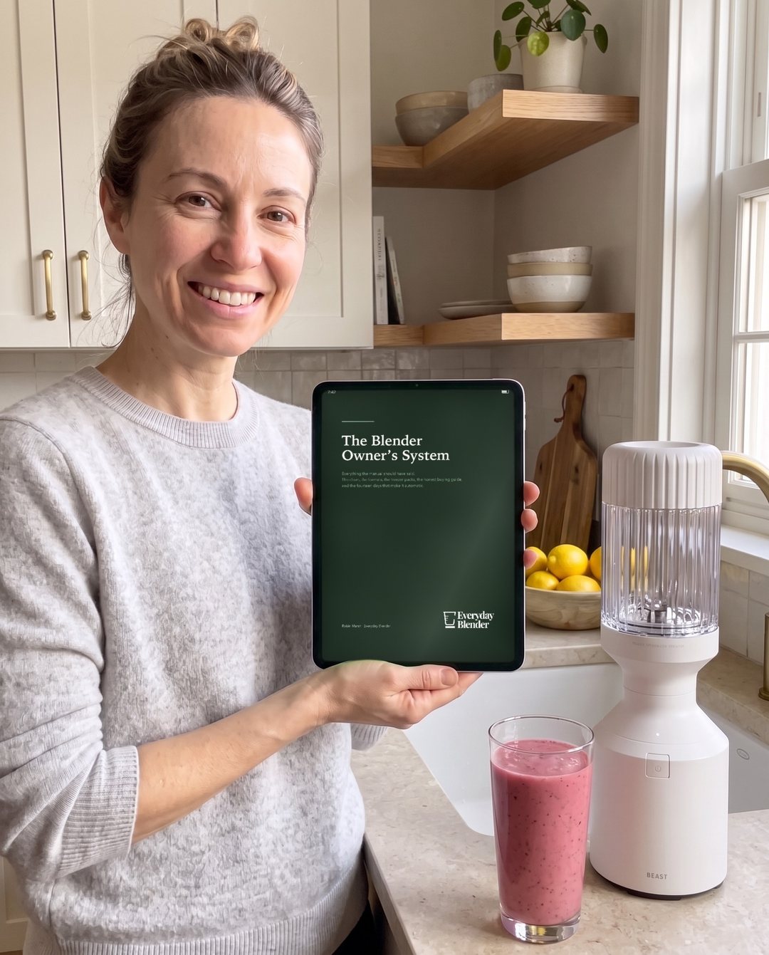 A woman in her kitchen holding up a tablet showing The Blender Owner's System, with a blender and a smoothie beside her