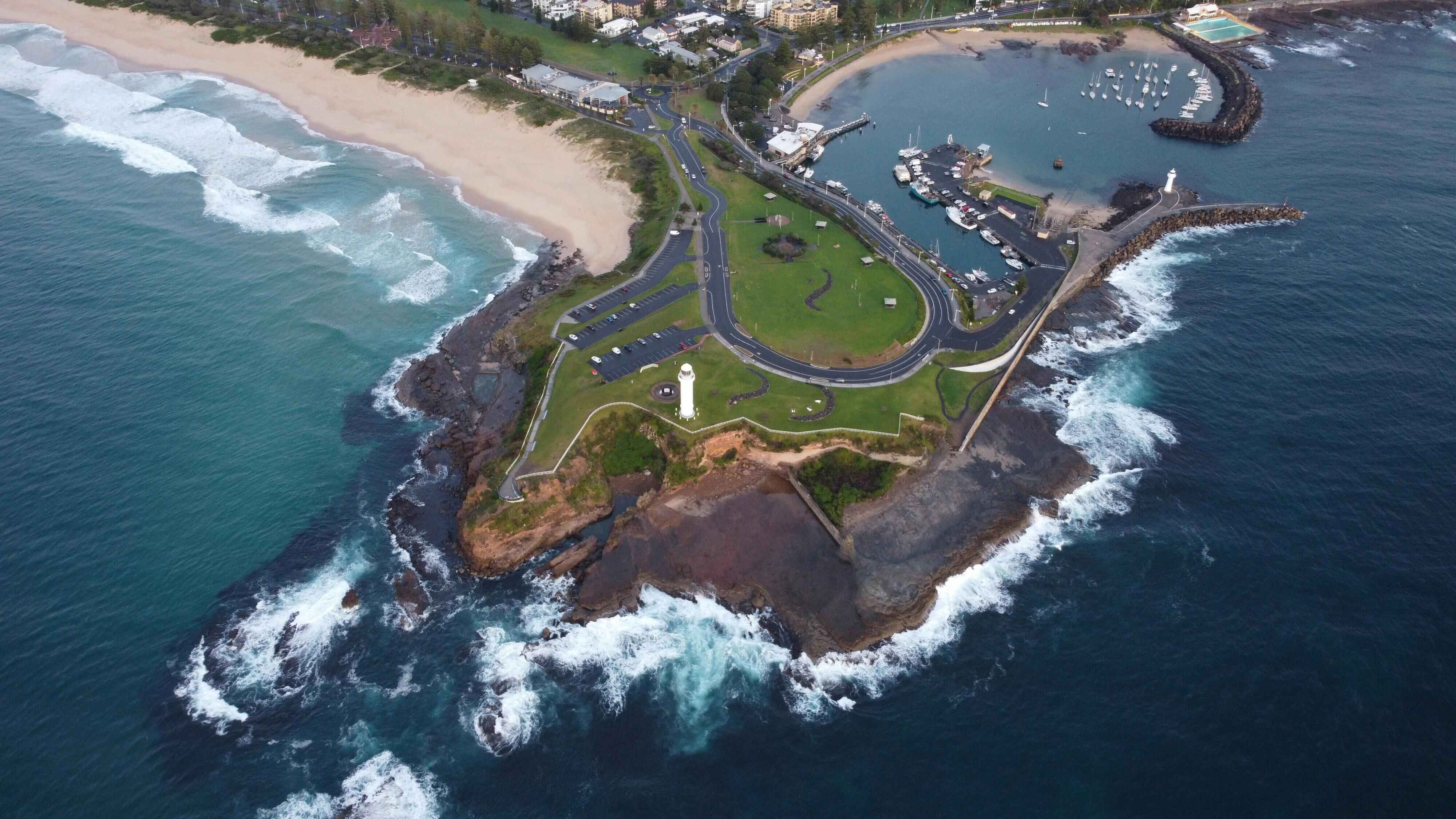 Aerial view of Wollongong coastal headland with beach, rocky shoreline, marina with boats, and a lighthouse surrounded by ocean waves. Aerial view of Wollongong coastal headland with beach, rocky shoreline, marina with boats, and a lighthouse surrounded by ocean waves.