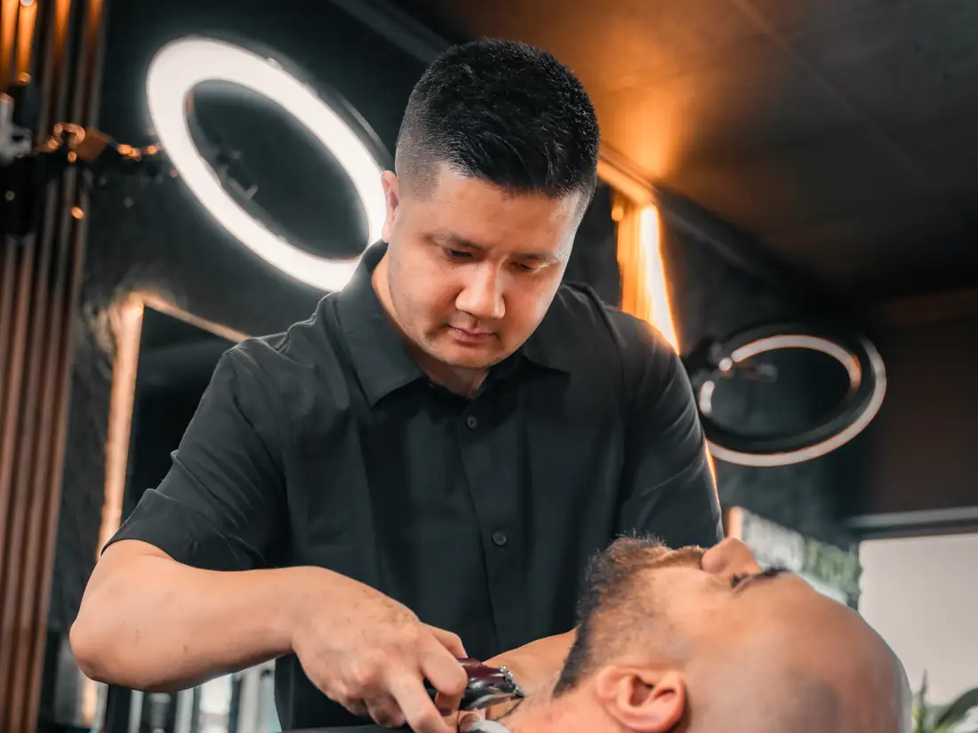 A barber using a straight razor along a reclined client&rsquo;s beard line