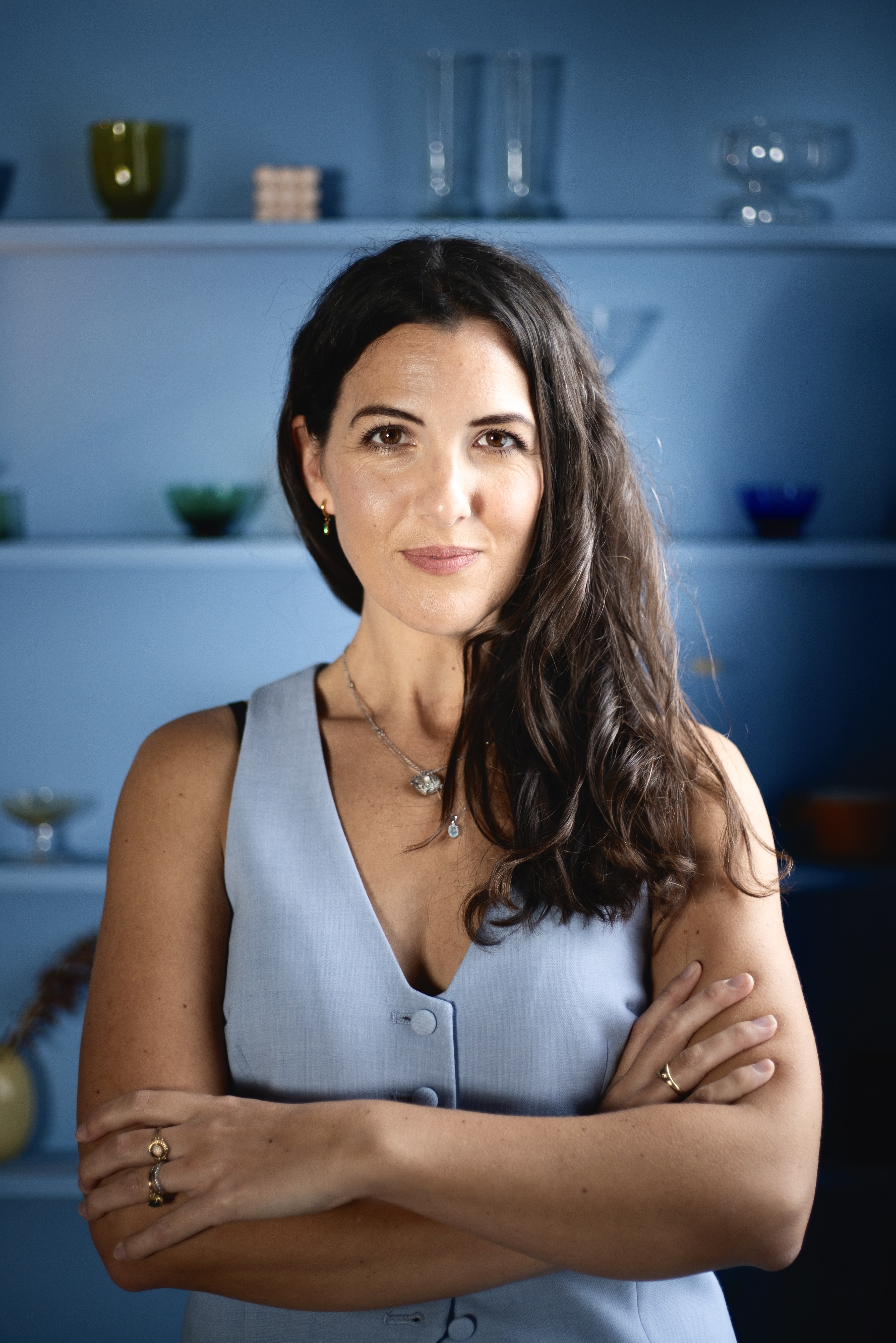 Alice Rose in a pale blue waistcoat, arms folded, in front of blue shelves