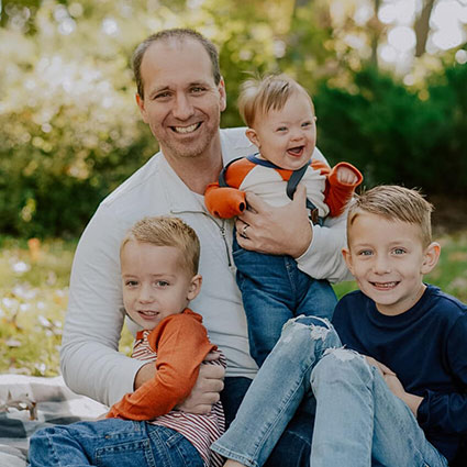 Young father with his 3 young sons, posing in the grass and smiling for the camera.