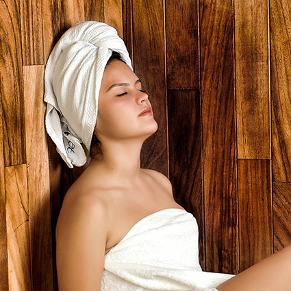 A young woman wearing a towel on her body and head, relaxes in the sauna with her eyes closed.