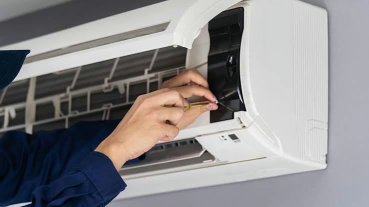 Technician in a blue uniform using a screwdriver to service a wall-mounted white air conditioning unit