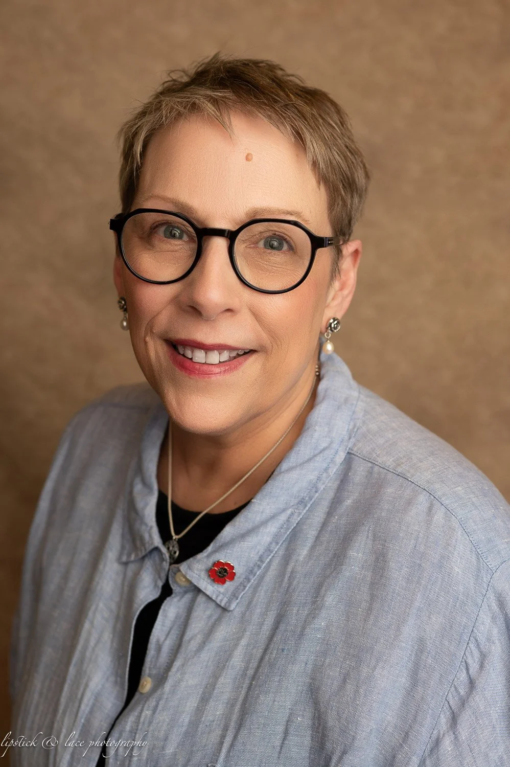 Person wearing glasses and a blue shirt with a poppy pin, smiling at the camera.