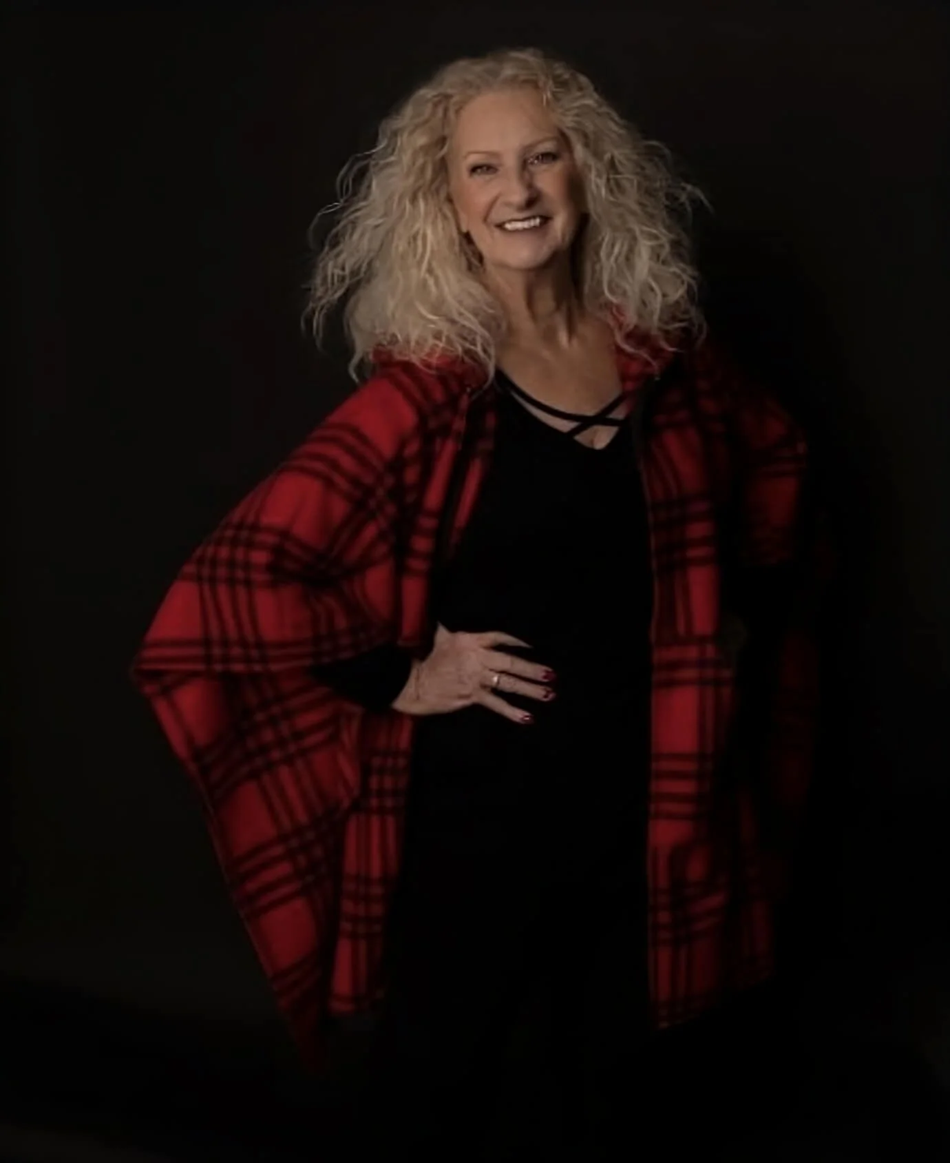 Person with curly blond hair wearing a red plaid poncho and black outfit, posing with a smile against a dark background.