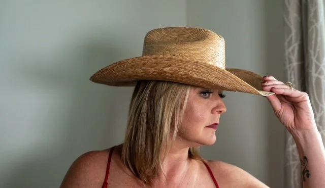 Woman in a cowboy hat looking to the side, touching the brim with her hand, wearing a red spaghetti strap top.