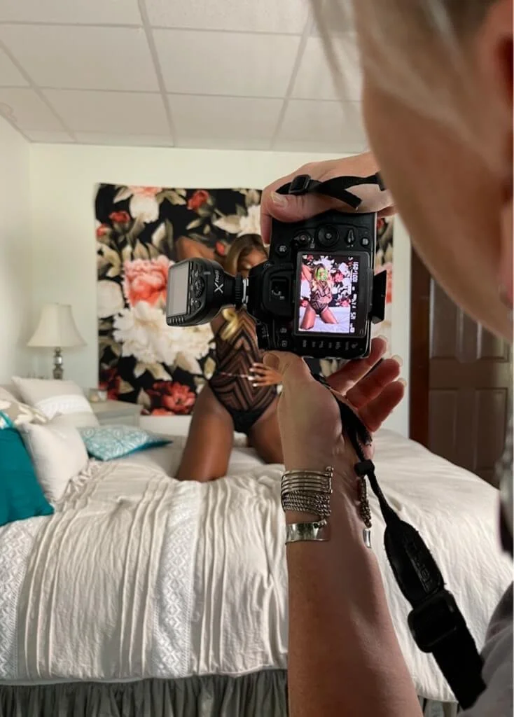 Photographer taking a picture of a model in a bedroom, with a camera displaying the preview image. The model poses on a bed with a floral backdrop.