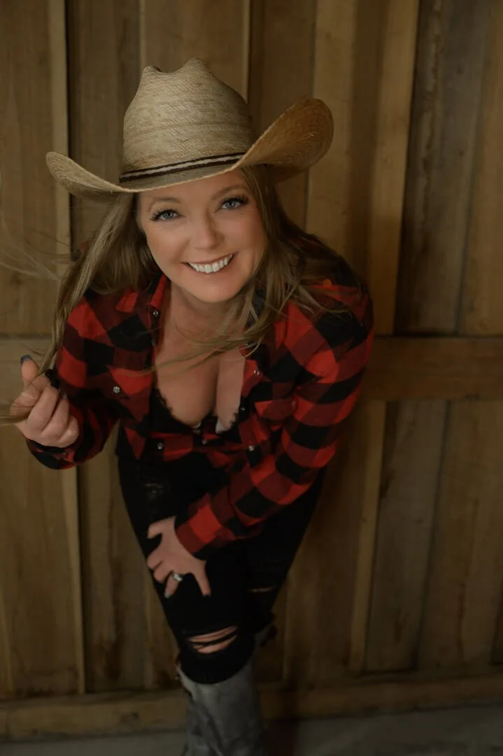 Smiling woman in a cowboy hat and red plaid shirt standing against a wooden background, holding a strand of her hair.