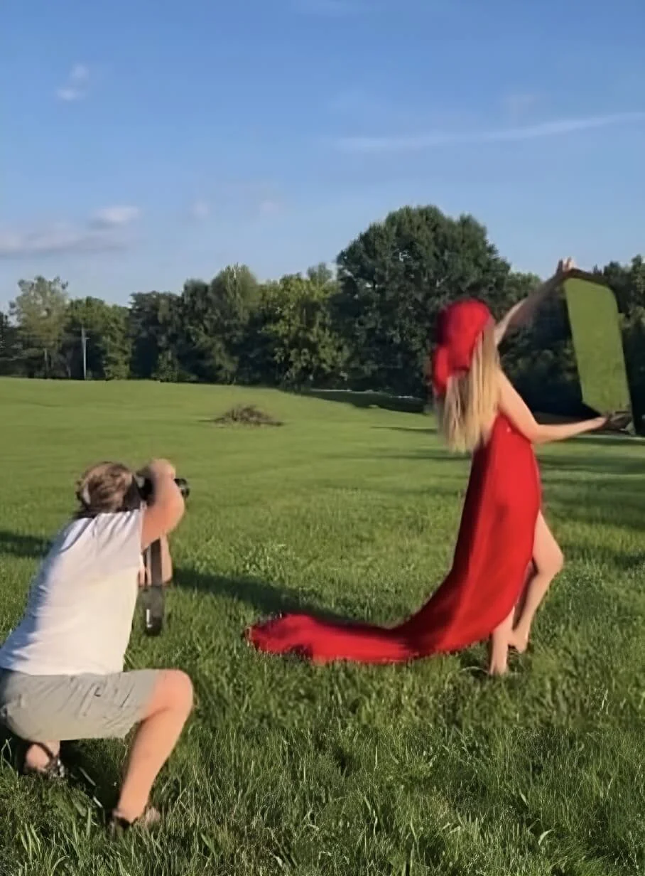 A photographer kneeling on grass takes a photo of a person in a flowing red dress holding a mirror, with trees and sky in the background.