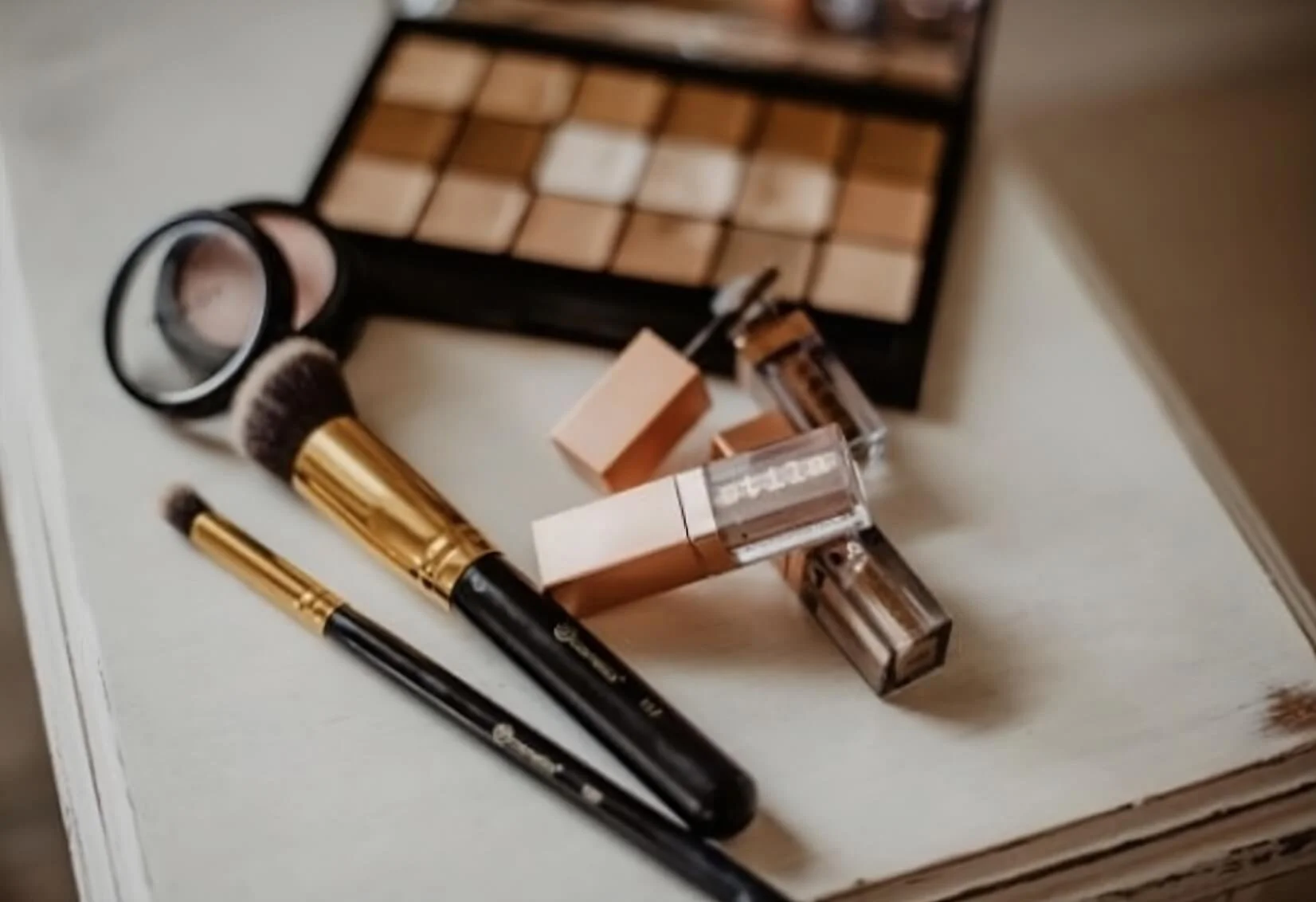 Makeup brushes, eye shadow palette, and lip gloss on table