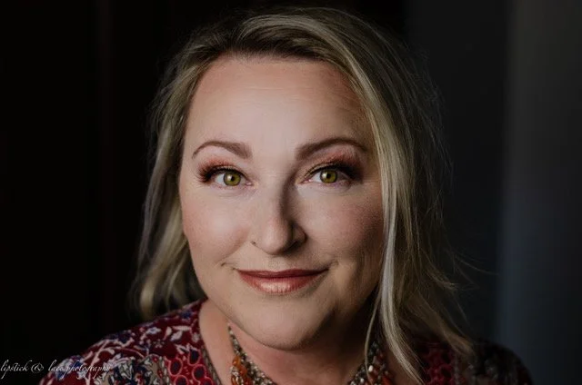 Portrait of a woman with blonde hair and light eye makeup, wearing a patterned blouse and a necklace.