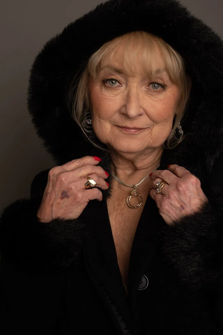 Older woman wearing a black fur hooded coat, holding the edges with her hands, displaying rings and a necklace.
