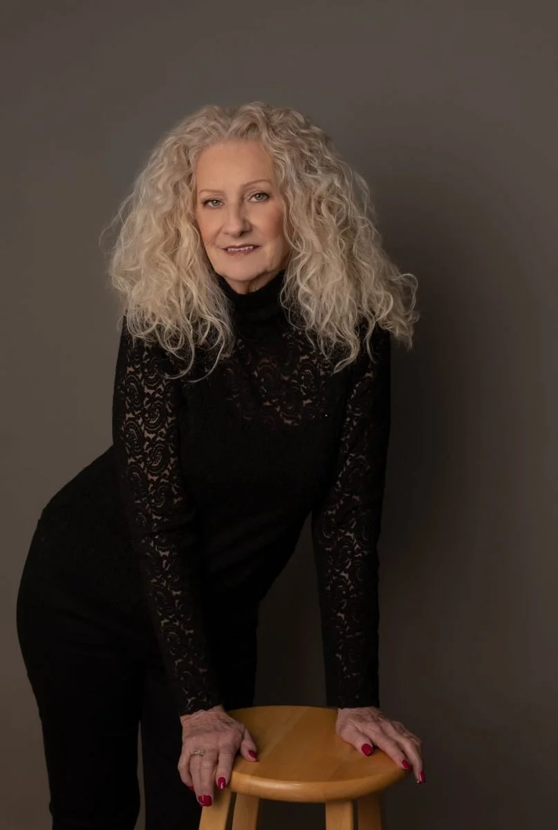 Woman with curly blond hair, wearing a black lace top, leaning on a wooden stool, against a gray background.