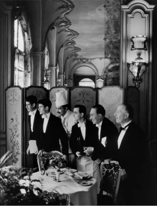 A vintage black-and-white photograph of waiters in tuxedos preparing a table