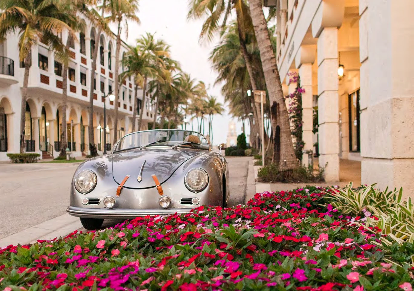 Classic silver convertible parked on a palm-lined Palm Beach street with flower beds