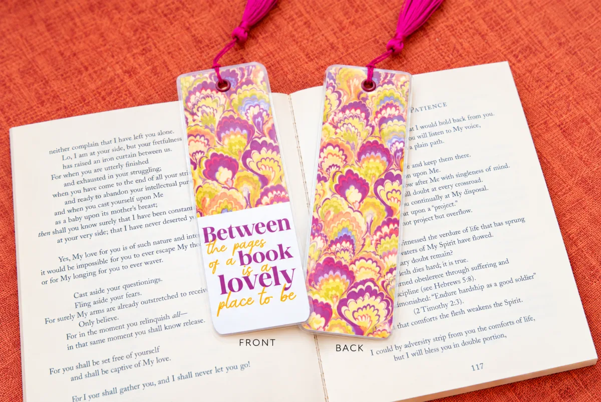 Double-sided laminated bookmark shown front and back, with a pink tassel resting on an open book