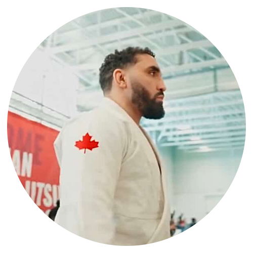 Coach Marcus, a tall, muscular man with a shaved head and sleeve tattoos, wearing a black rash guard, stands confidently in front of a wall of championship medals. The background is a bright, clean gym with blue mats.