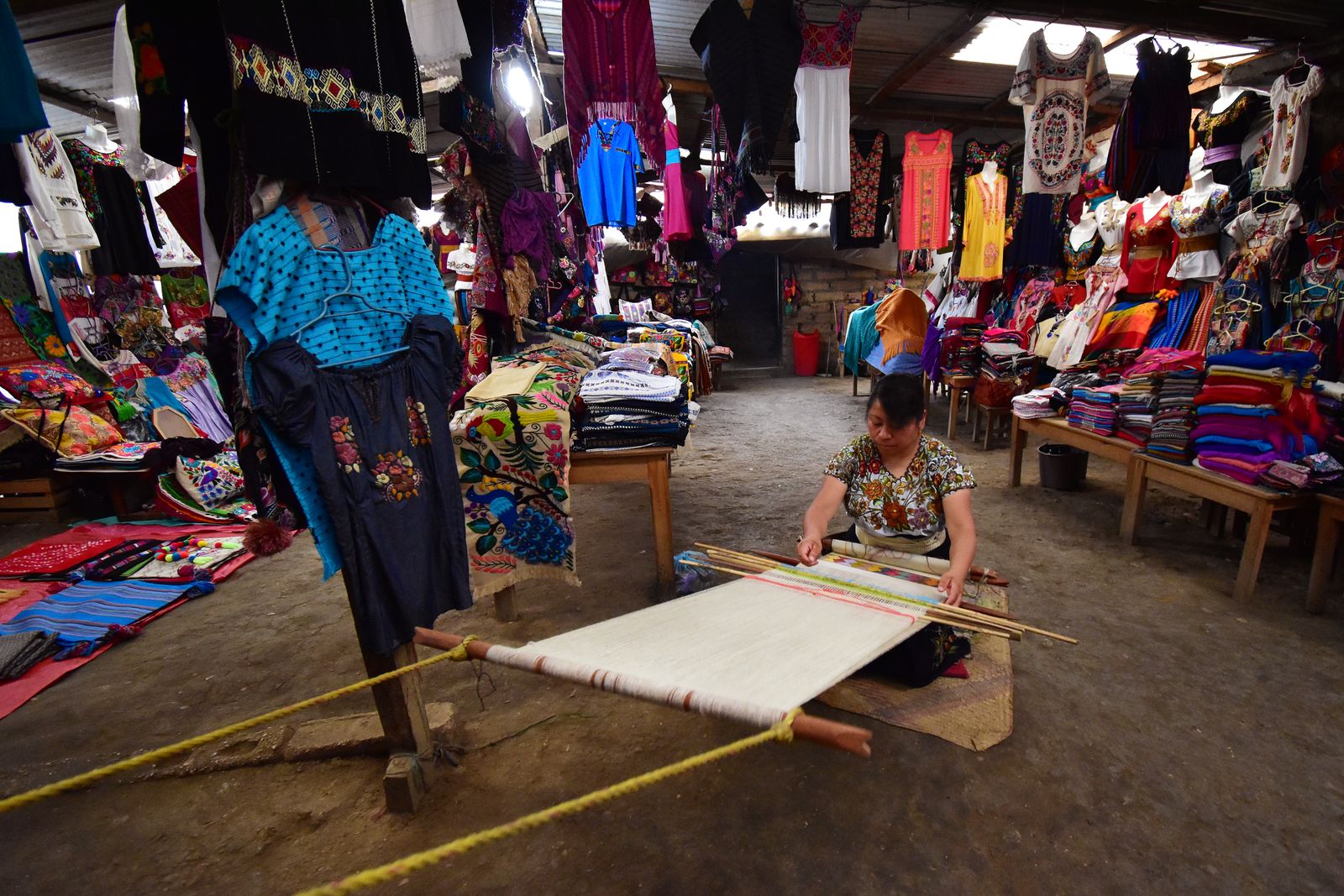 A Zinacant&aacute;n weaver working a backstrap loom on the floor of a textile co-op shop hung with embroidered blouses