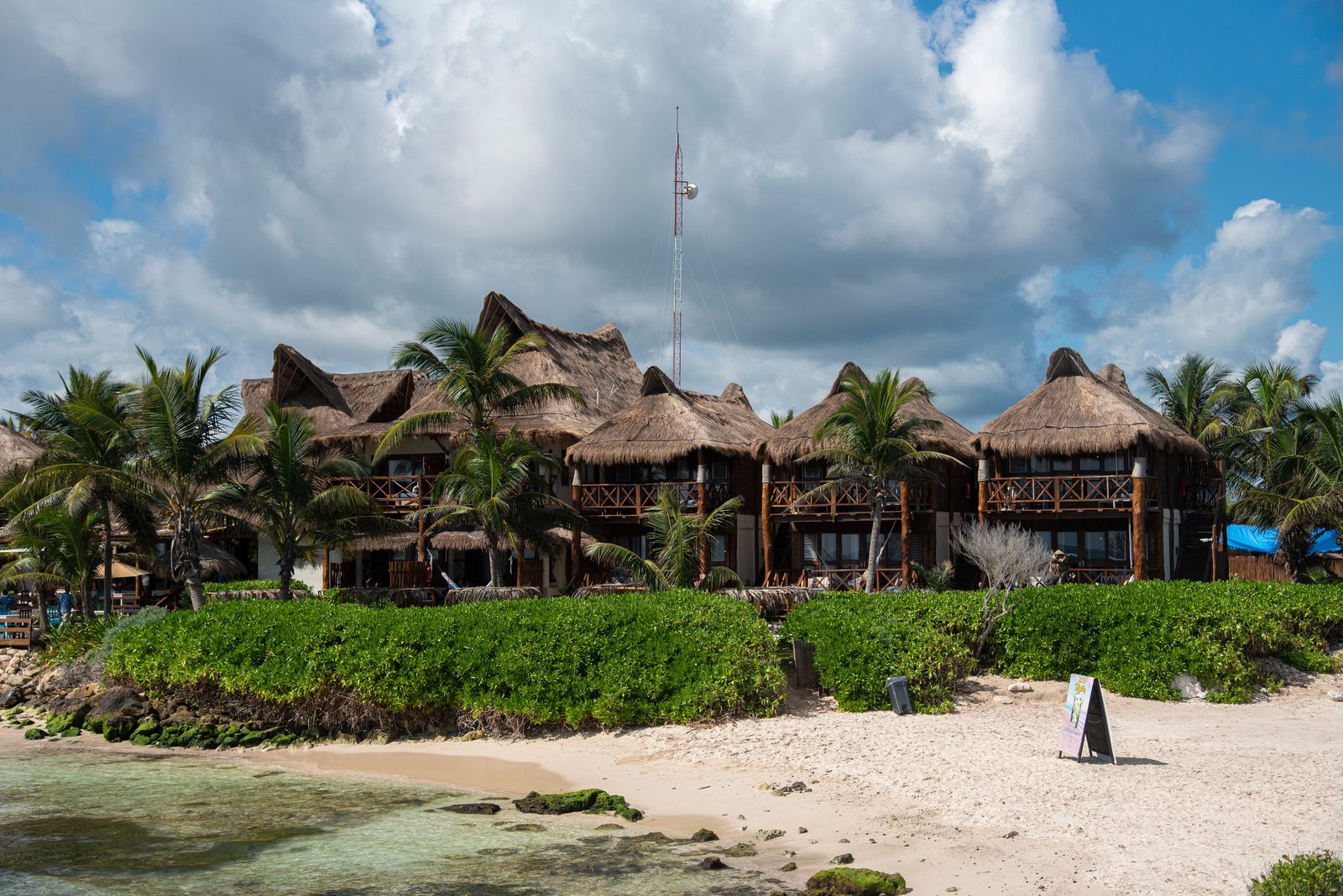 A row of eco-chic palapa-roofed hotels along Tulum's Zona Hotelera beach road, with the Caribbean visible between the properties