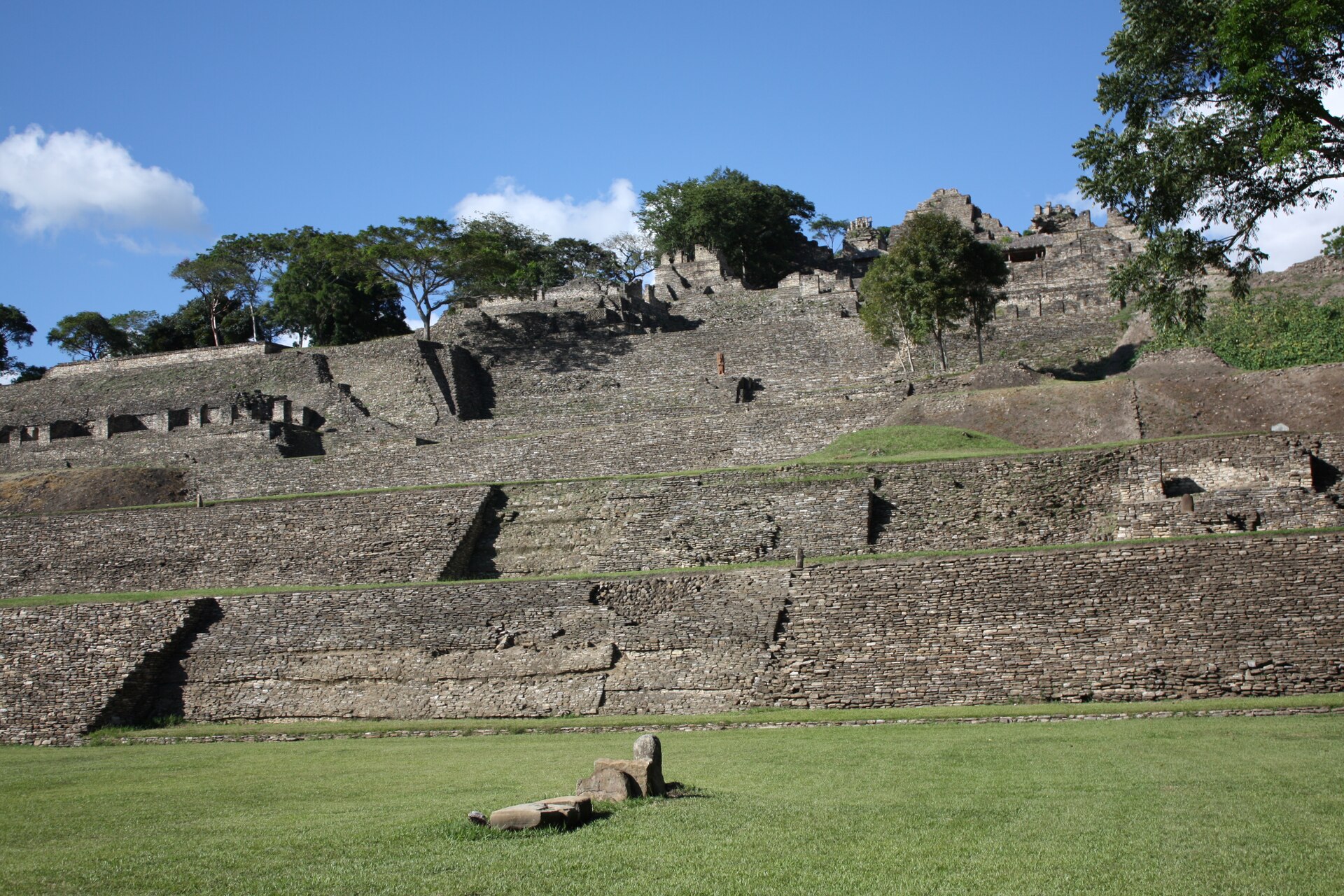 The terraced stone acropolis of Toniná climbing a green hillside above the plaza, its platforms and temples stacked under a bright sky The terraced stone acropolis of Toniná climbing a green hillside above the plaza, its platforms and temples stacked under a bright sky