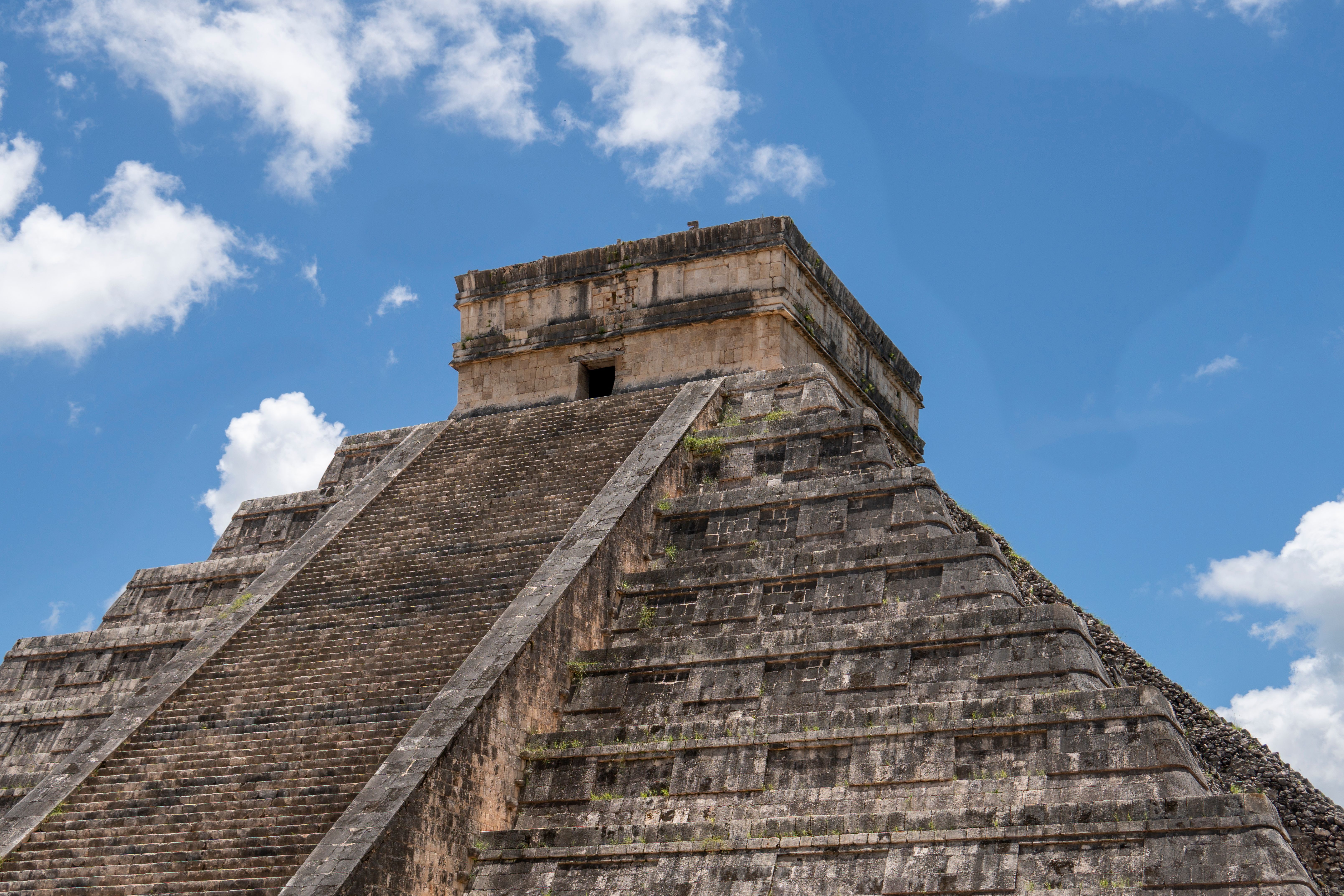 A wide shot of El Castillo, the main step-pyramid at Chichén Itzá, under hard midday light with tour groups gathered on the grass in front of it.