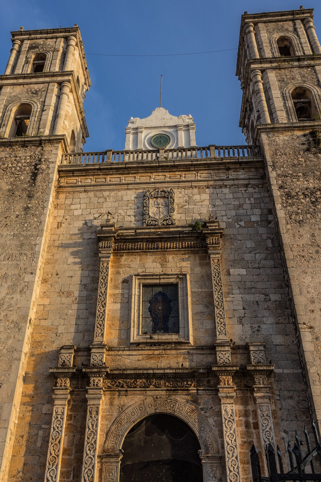 A colonial plaza in Valladolid at golden hour, pastel single-story buildings around a church, a few people walking, no crowds
