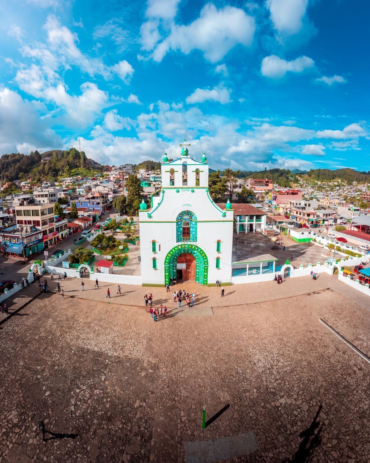 Aerial view of the white-and-green church of San Juan Chamula on its wide stone plaza, with the town and green hills behind