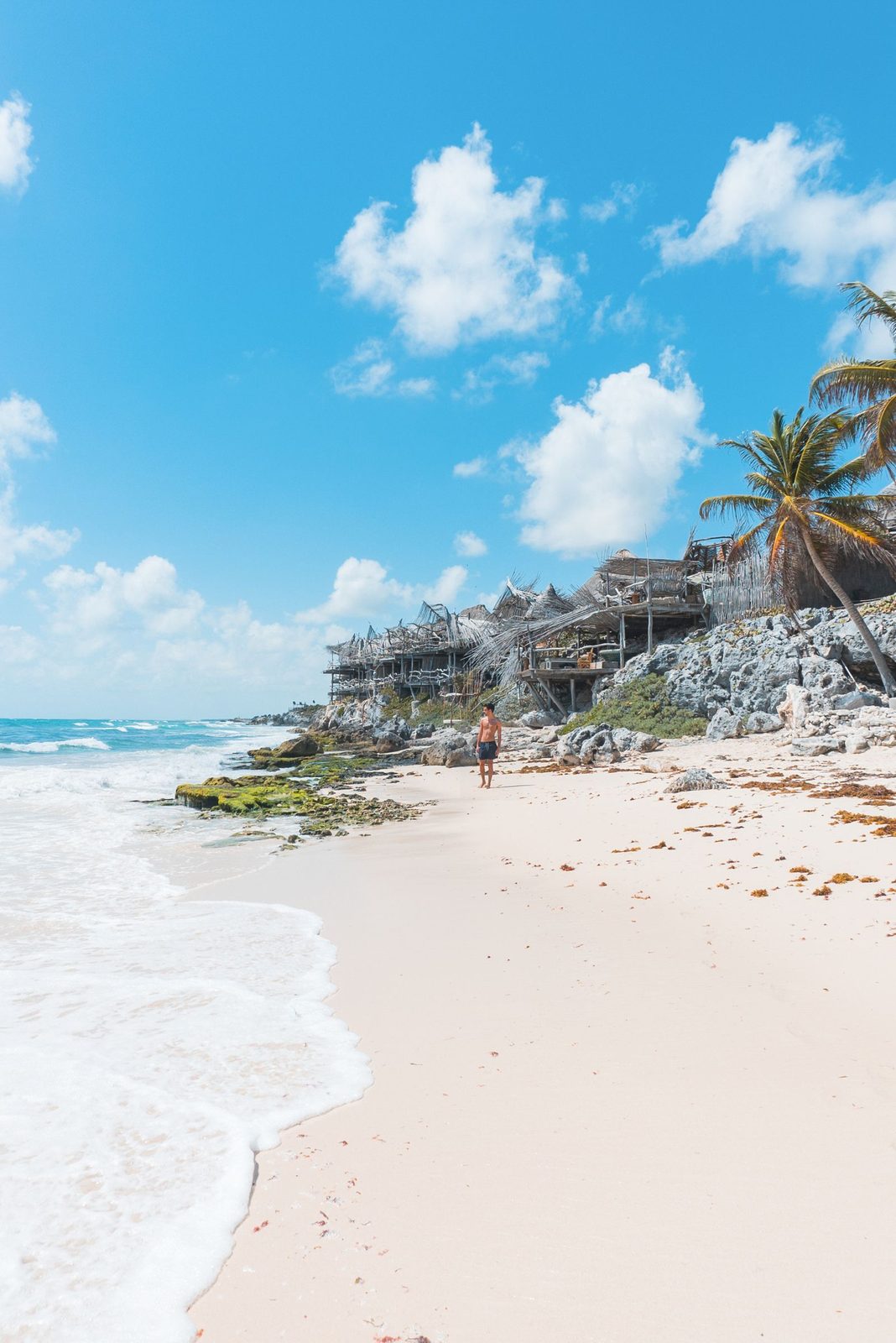 Tulum's white-sand beach with rocky shore and palms under a bright sky