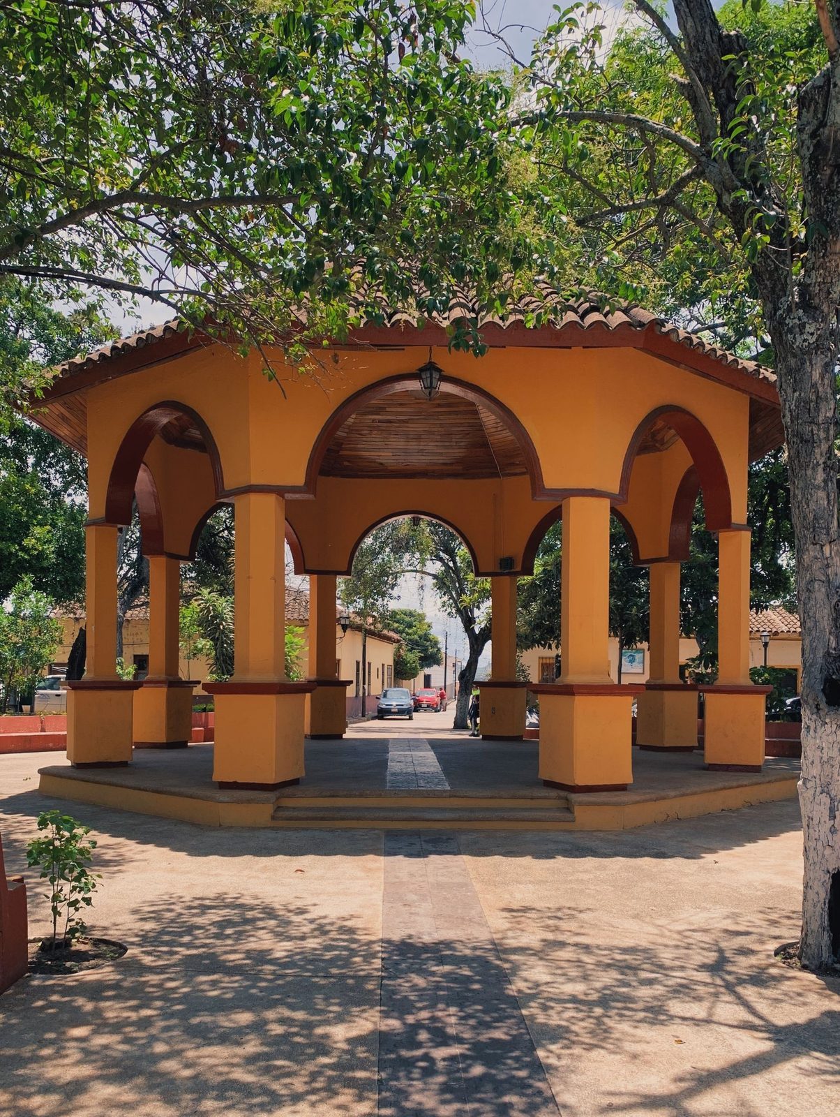 A yellow arched gazebo in a tree-shaded park in Comitán de Domínguez