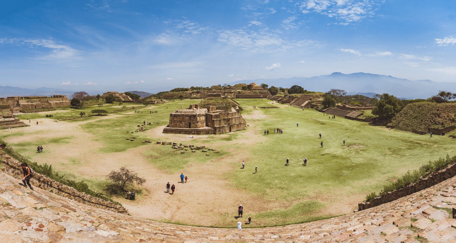 Panoramic view across the Great Plaza at Monte Albán, with its central temple platforms, visitors on the grass and the mountains of the Oaxaca Valley behind