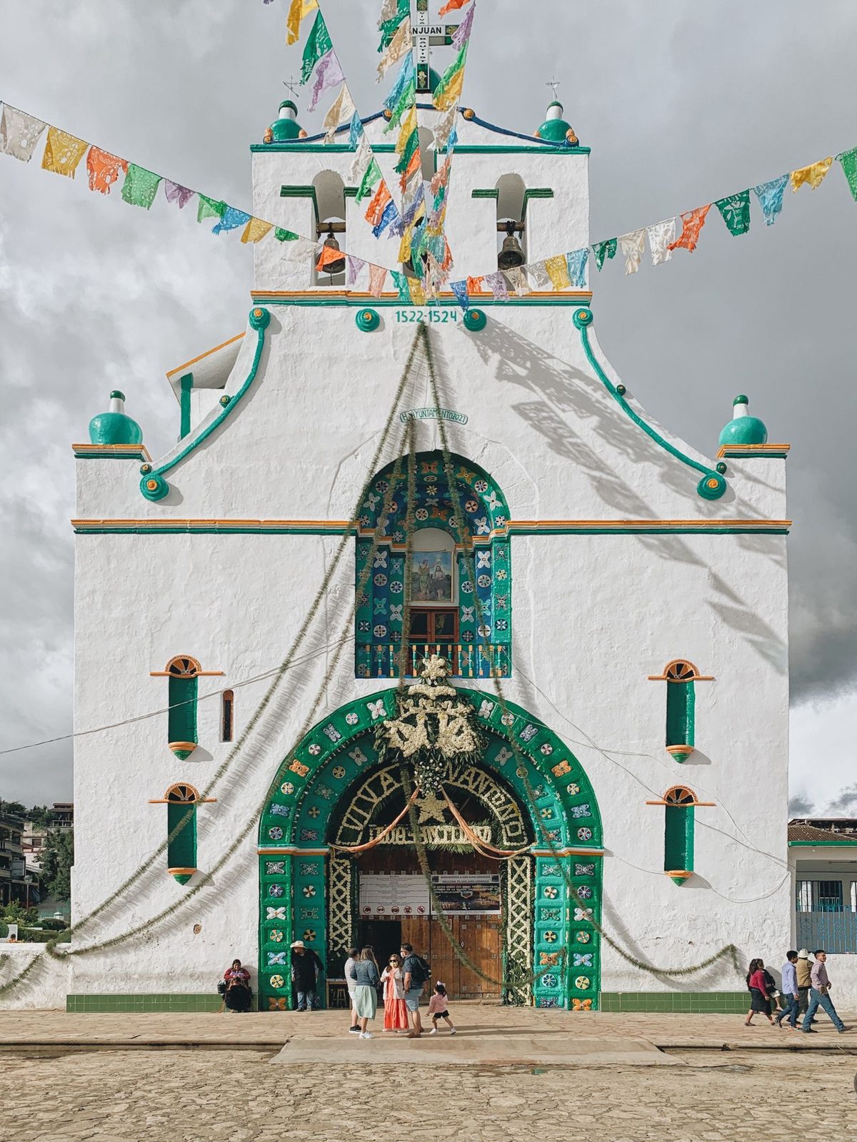 The whitewashed facade of the church of San Juan Bautista in San Juan Chamula, its arched portal tiled in green and blue under festival flags