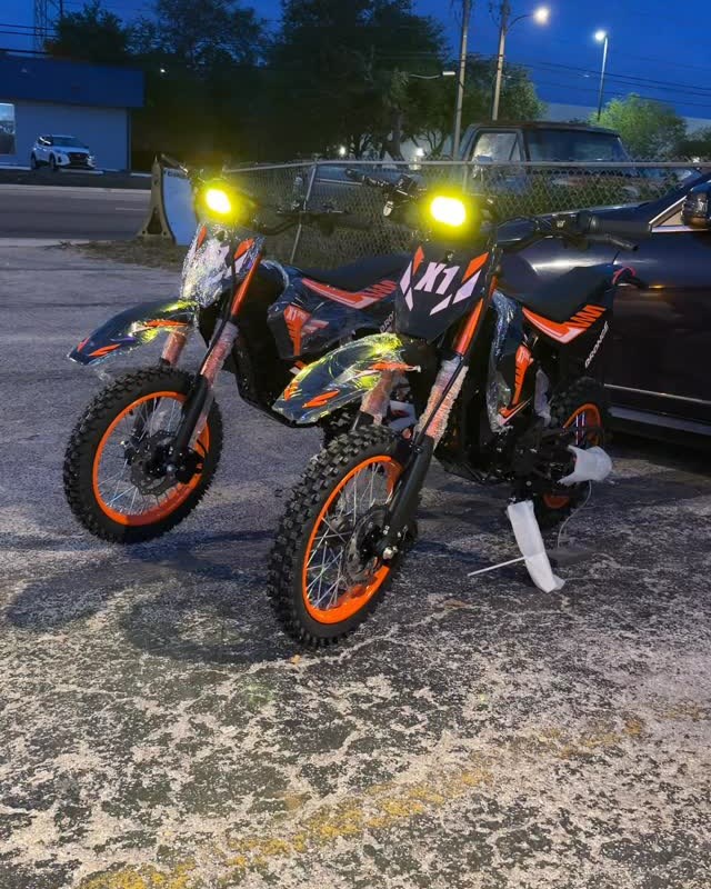 Two orange and black electric dirt bikes on the lot at dusk, headlights on.