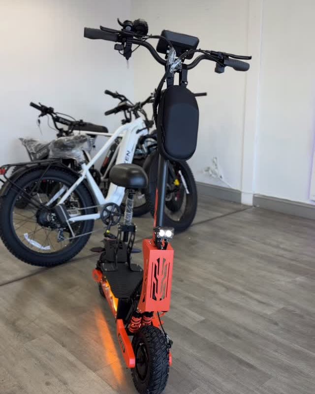 An orange electric scooter on the shop's wood floor, with two ebikes behind it.