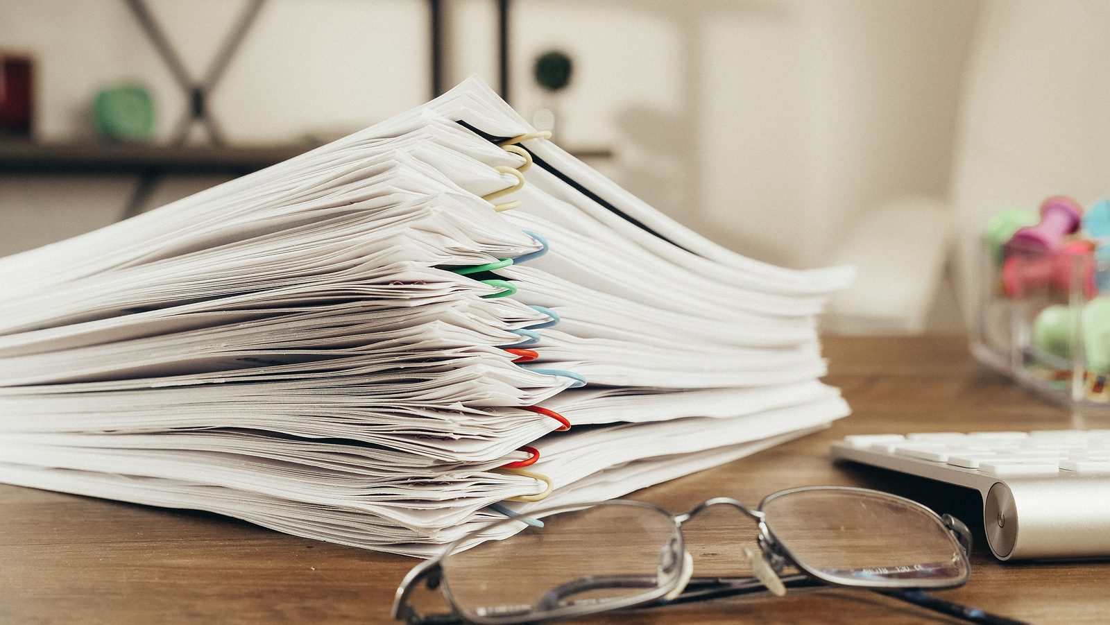 A tall stack of paper-clipped documents on an office desk beside a pair of glasses