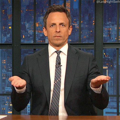 Male television host in a dark suit seated at a late-night talk show desk, holding both hands up with a puzzled expression. Male television host in a dark suit seated at a late-night talk show desk, holding both hands up with a puzzled expression.