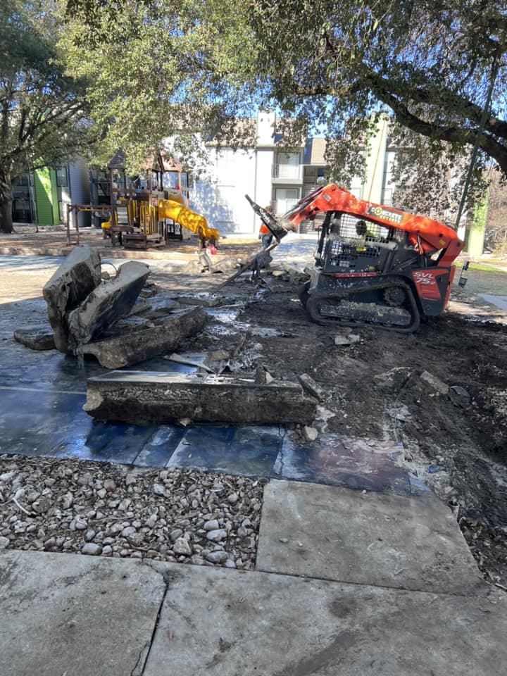 A concrete demolition crew breaking up a cracked residential driveway slab in San Antonio, Texas, with rebar exposed and broken concrete pieces staged for haul-off. The surrounding lawn and curb remain undisturbed.