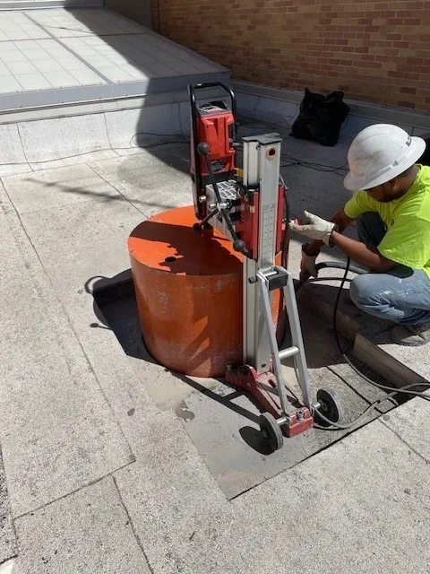 A concrete coring drill mounted on a slab floor inside a San Antonio commercial building, with a clean round hole drilled through the concrete surface. Dust control equipment is visible beside the drill rig.