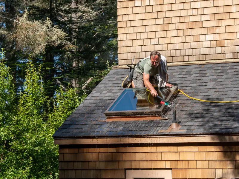 Worker cleaning skylight and shingle roof with hose.