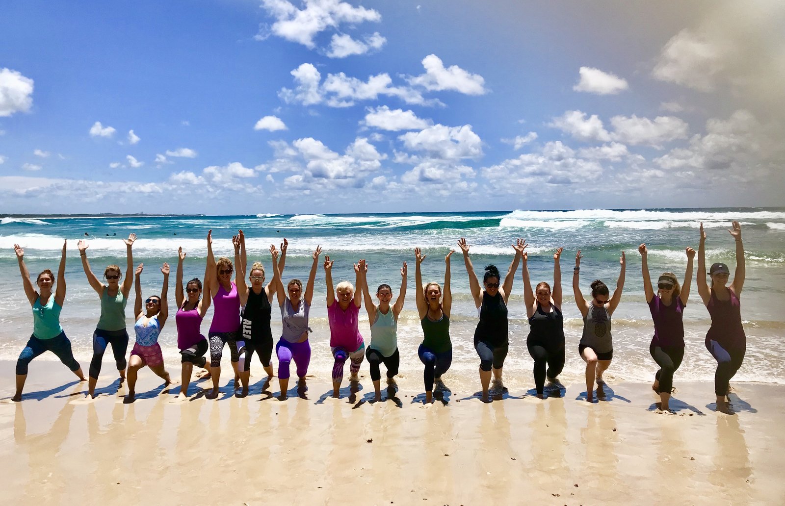 A group of women in yoga poses on the beach, arms raised to the sky