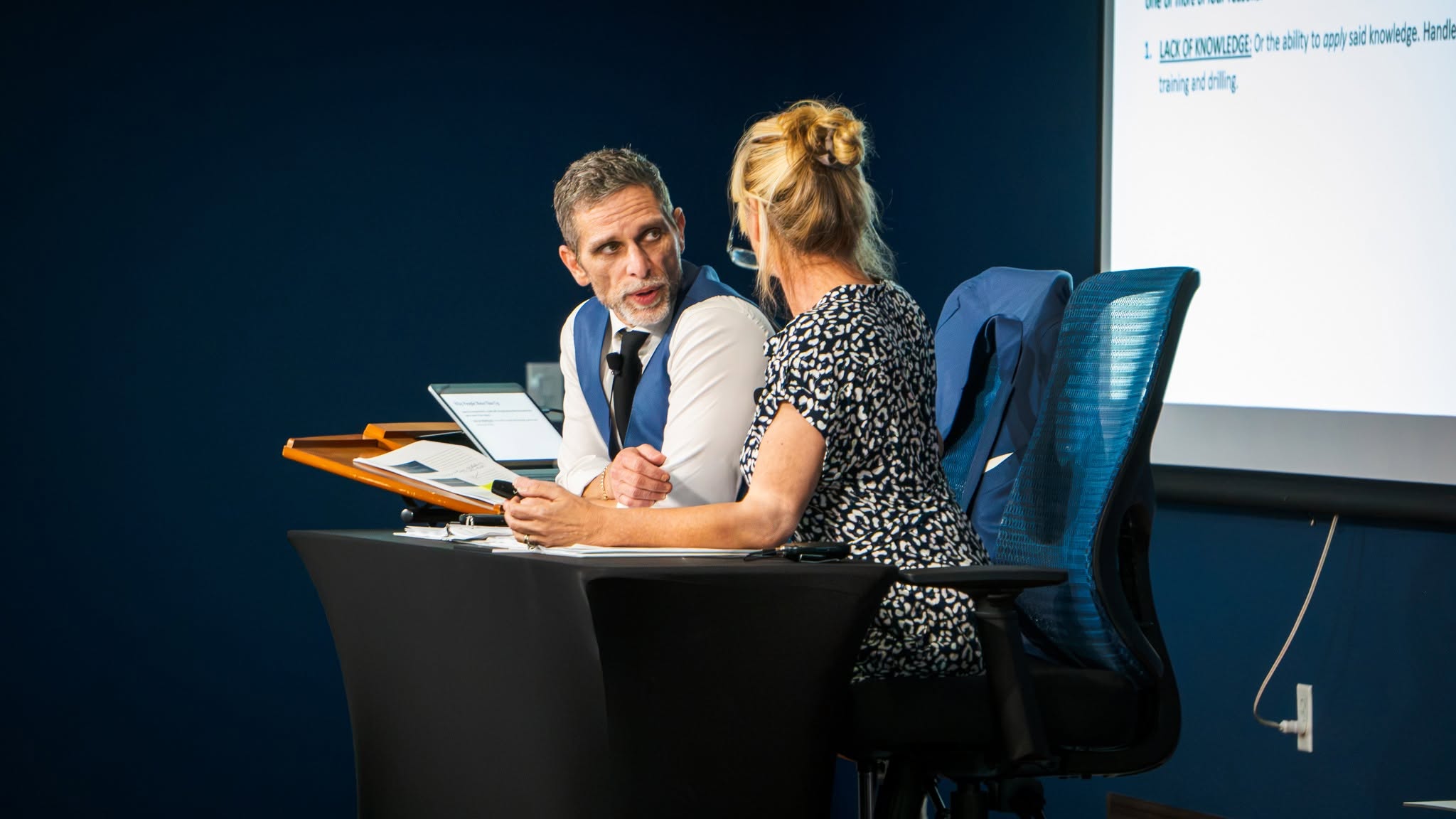 The two presenters conferring at the lectern beside a projected training slide
