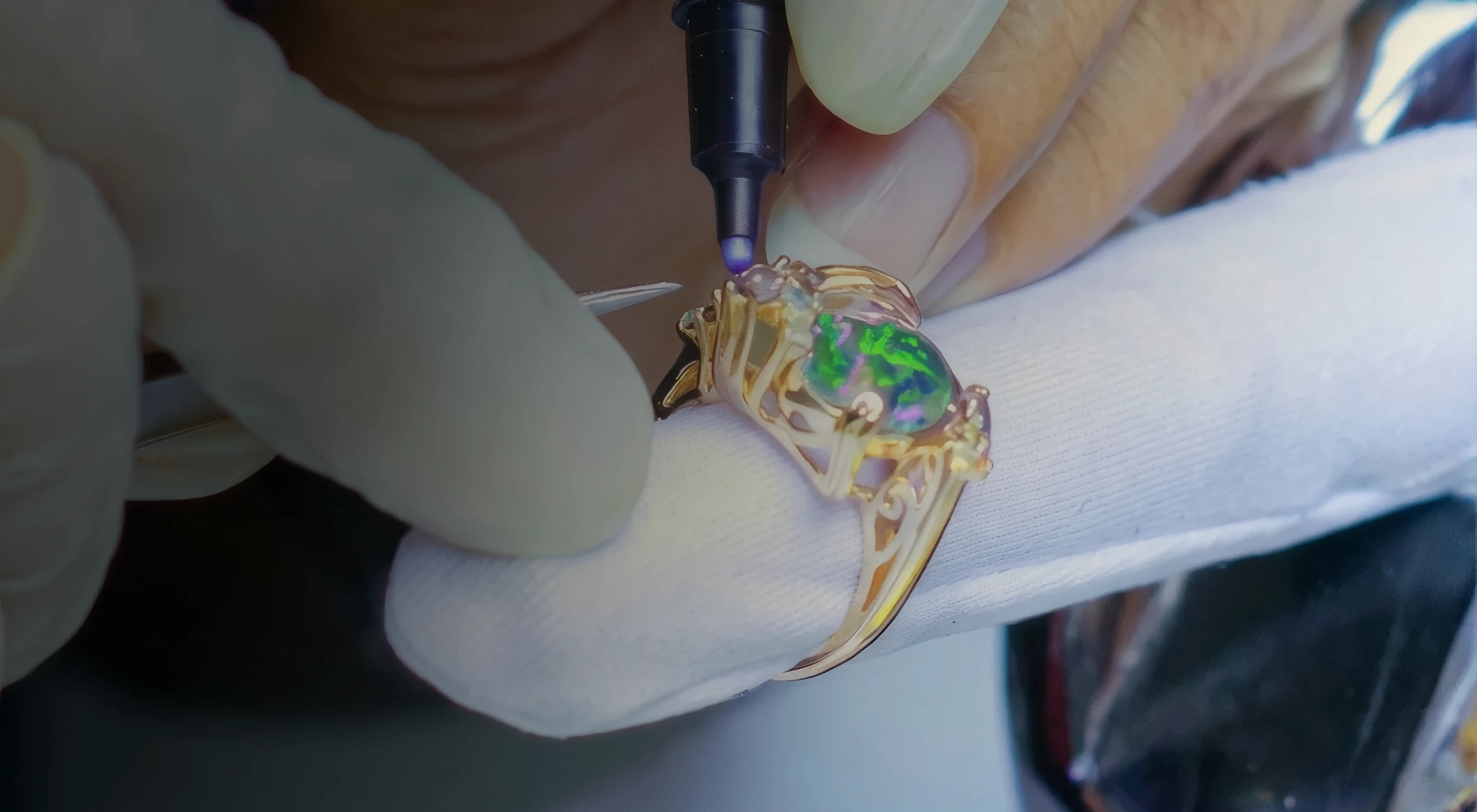 Gloved hands setting stones into a filigree gold opal ring at the bench