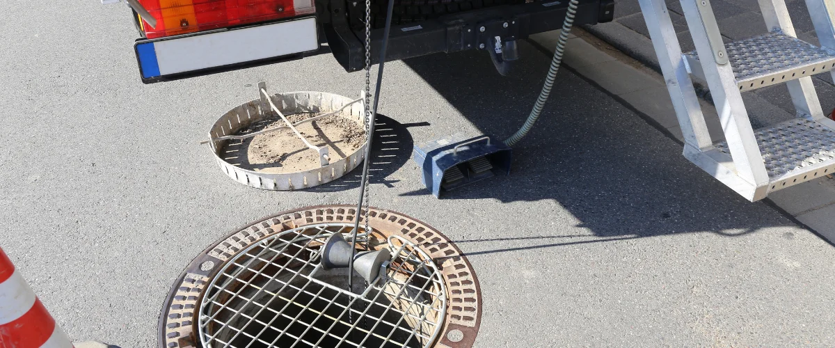 Sewer repair equipment next to an open manhole on asphalt, showing a chain with tools and a removed grate.